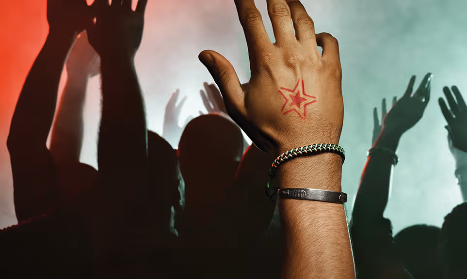 Raised hand with star tattoo and bracelets in front of a crowd with hands raised at a smoky event with red and green lighting.