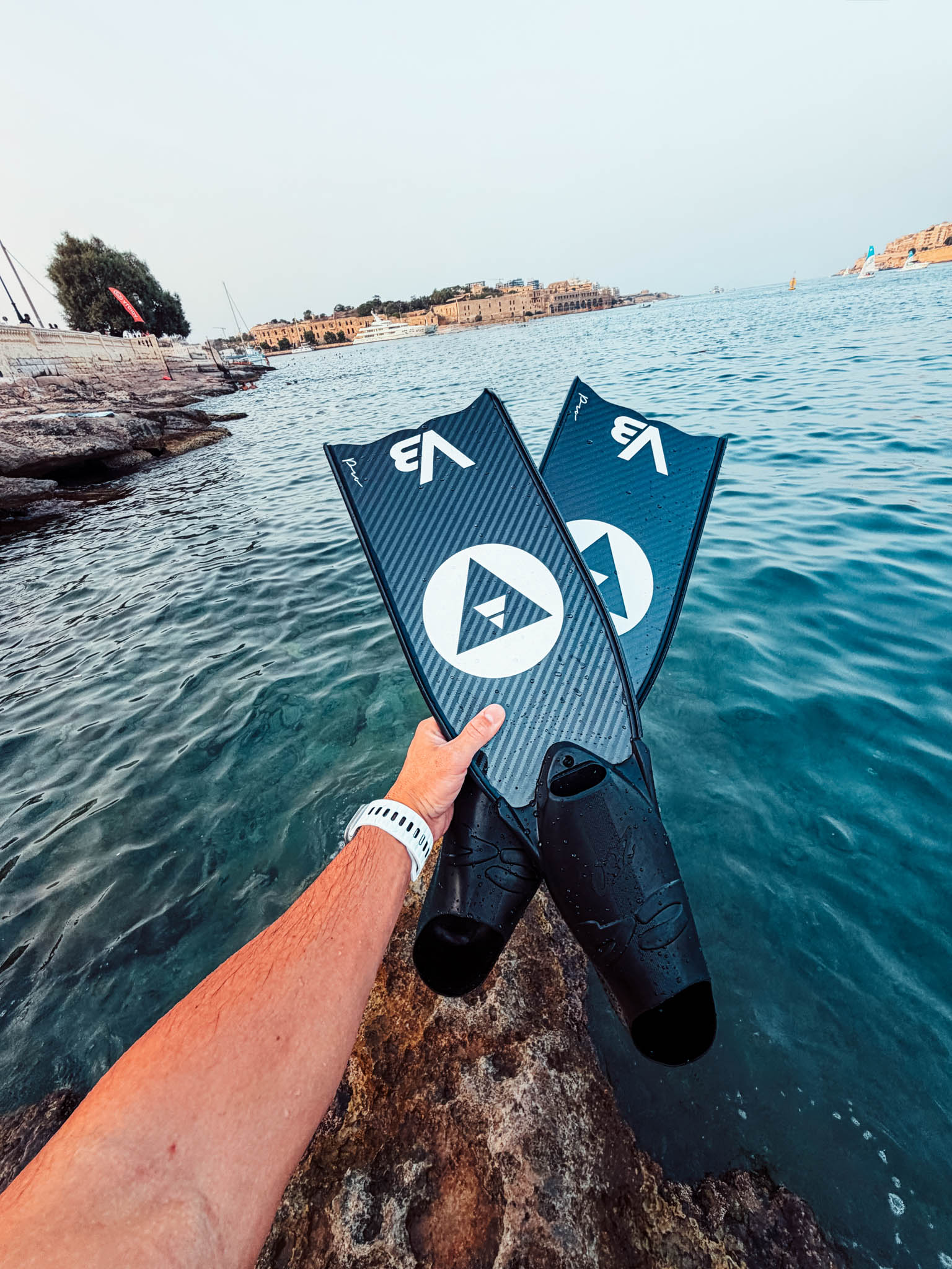 Hand holding a pair of Alchemy V3-30 Pro carbon fins above clear sea water