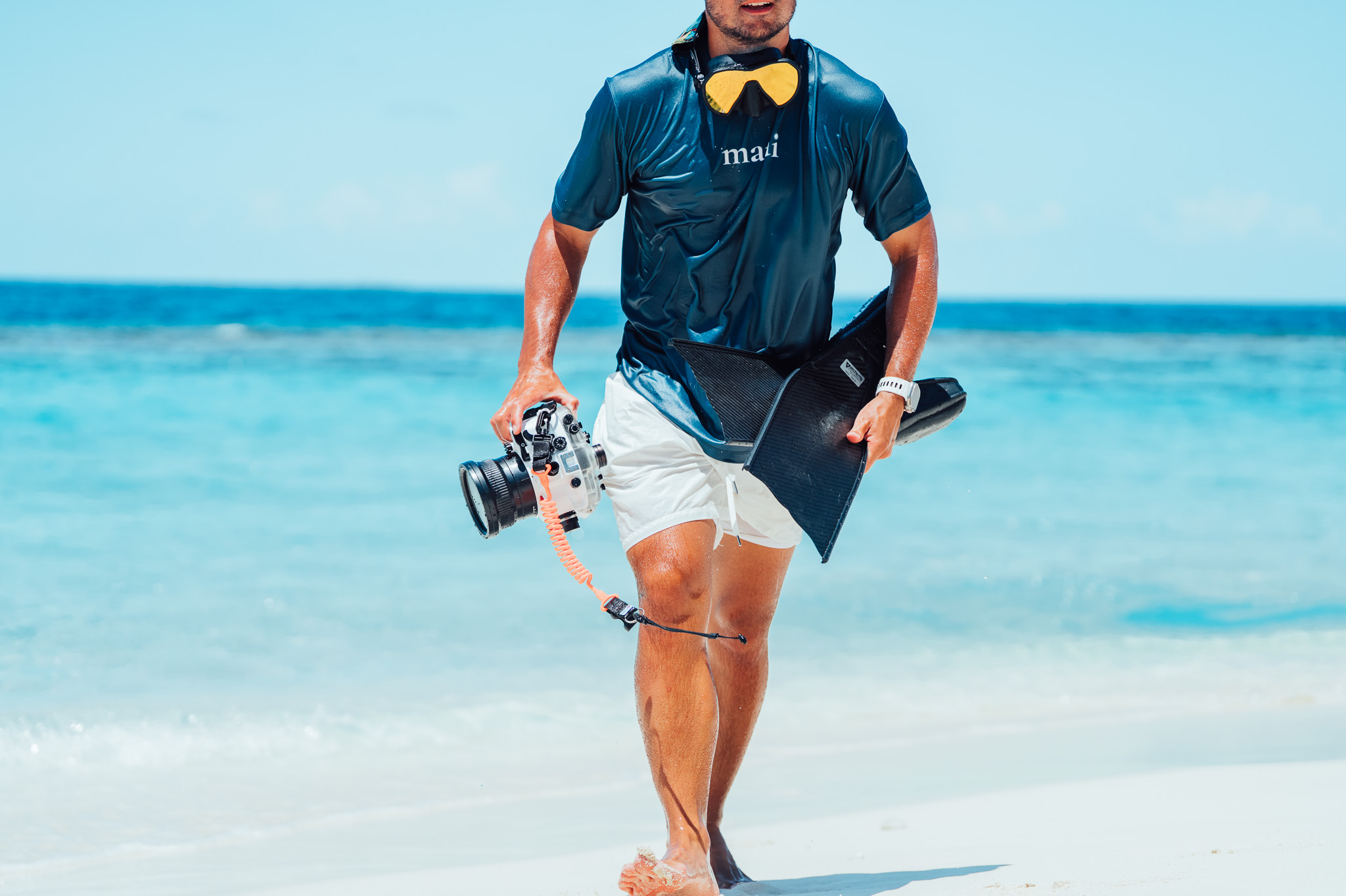 Snorkeler walking up the beach with a gold mirrored mask around his neck, carbon fins and a camera