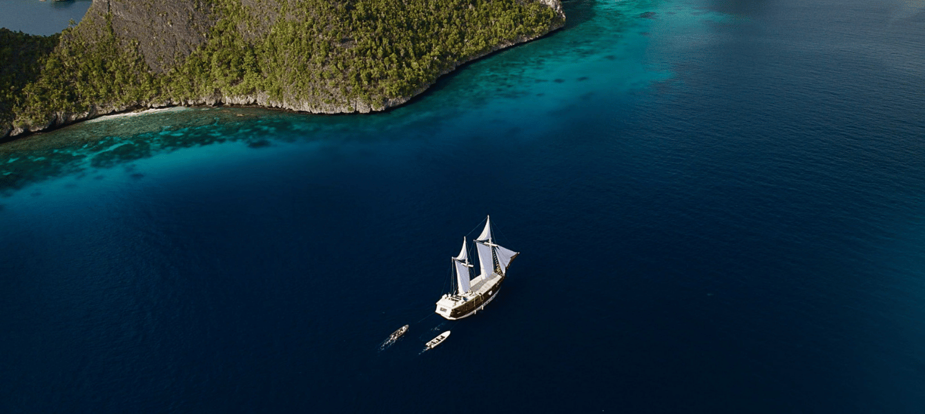 Aerial view of a sailing liveaboard anchored off a jungle island in Raja Ampat