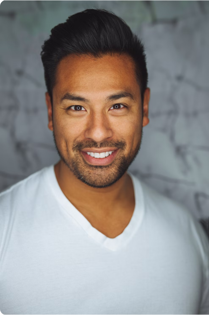 Smiling man with dark hair and beard wearing a white V-neck shirt in front of a blurred background.