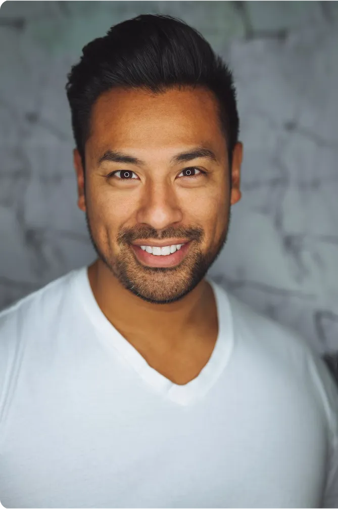 Smiling man with dark hair and beard wearing a white V-neck shirt in front of a blurred background.