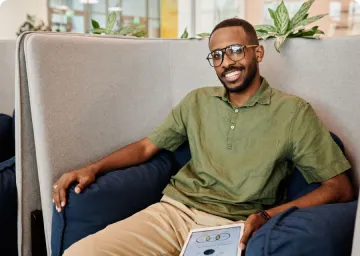 Smiling man with glasses sitting on blue armchair with green plants and window in the background.