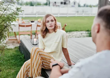 Woman in yellow shirt and striped pants sitting outdoors, talking to a man facing her.
