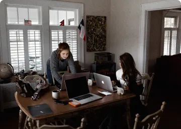 Two women working with laptops at a wooden table in a room with windows and flags.