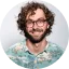 Smiling man with curly hair, beard, and glasses wearing a denim shirt against a neutral background.