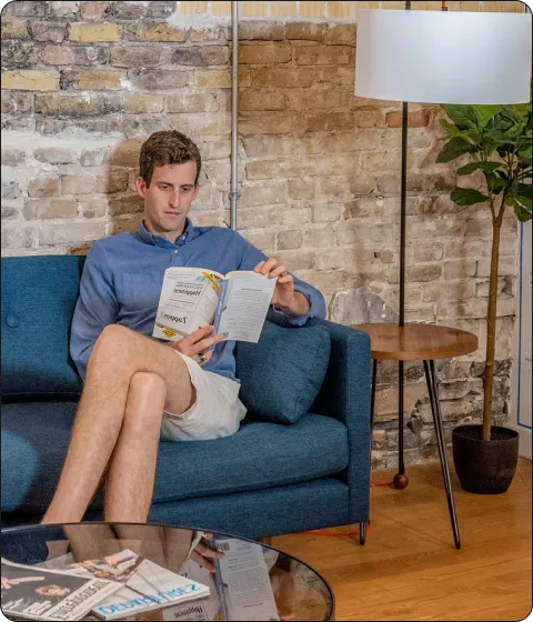 Man in a blue shirt and shorts sitting on a blue couch reading a book in a modern room.