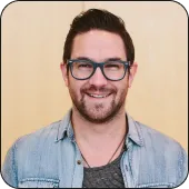 Smiling man with glasses, beard, and casual denim shirt against a light wooden background.