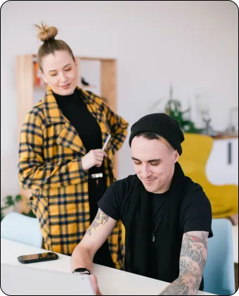 Woman in yellow plaid coat watches man with tattoos working on a laptop at a desk.