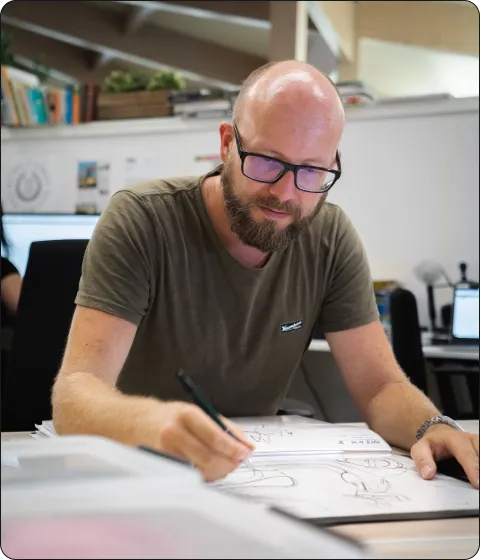 Bald man with glasses drawing sketches on paper at a desk in an office setting.