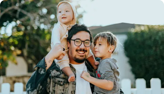 Man with glasses carrying a baby on his shoulders and holding a toddler close outdoors near a white picket fence.