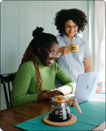Two women smiling and working on a laptop at a table with a coffee carafe.