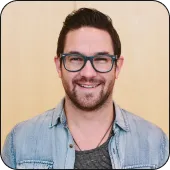 Smiling man with glasses and beard wearing a denim jacket in front of a wooden background.