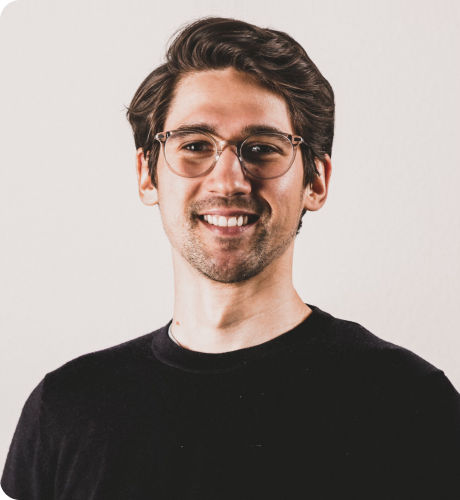 Smiling young man with glasses and dark hair wearing a black shirt against a plain light background.