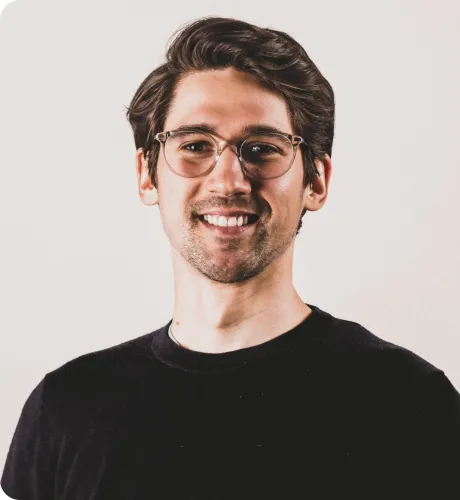 Smiling young man with glasses and dark hair wearing a black shirt against a plain light background.