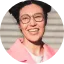 Smiling young person wearing glasses and an orange shirt in front of a wall.