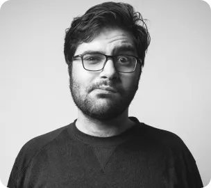 Bearded man wearing glasses and a dark sweater looking skeptically with one eyebrow raised.