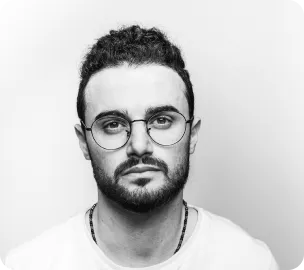 Black and white photo of a serious man with glasses, curly hair, beard, and a white t-shirt.