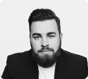 Bearded man with dark hair wearing a black jacket and white shirt, looking at the camera.