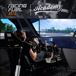 Person sitting in a racing simulator setup with steering wheel and pedals, surrounded by large screens showing a racetrack.