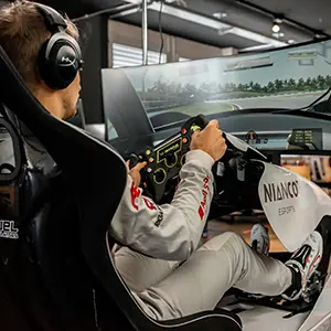 Man in racing suit and headset using a steering wheel controller to race on a simulator with multiple screens.