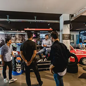 Four men standing and talking in an indoor automotive showroom with cars and tires in the background.