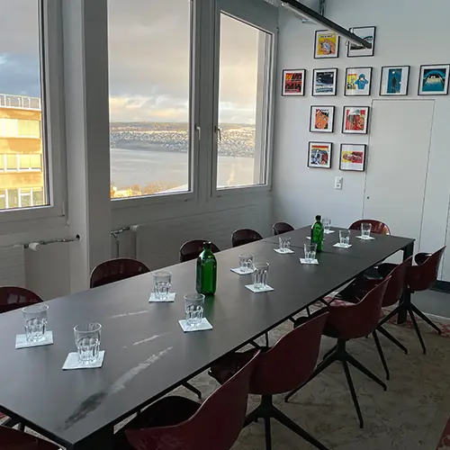Modern conference room with a long black table, ten maroon chairs, glasses on coasters, two green bottles, and framed artwork on the wall, overlooking a large window with a river view.