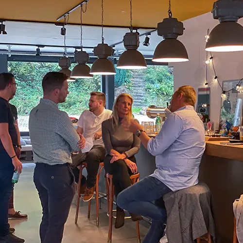Five adults socializing in a cozy indoor setting with large windows, pendant lighting, and a wooden bar counter.