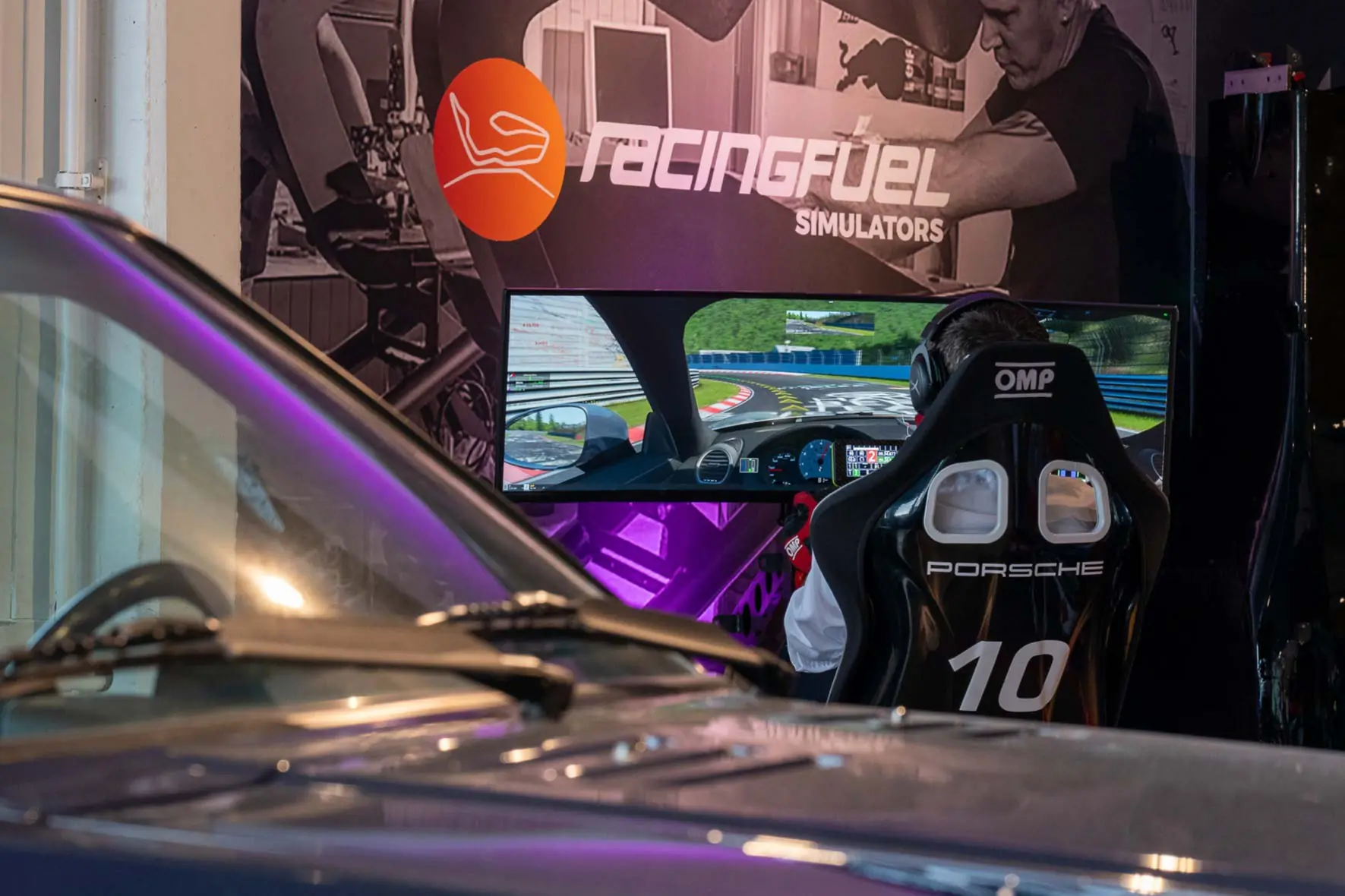 Person seated in a Porsche racing simulator chair facing an ultra-wide screen displaying a racing game track.