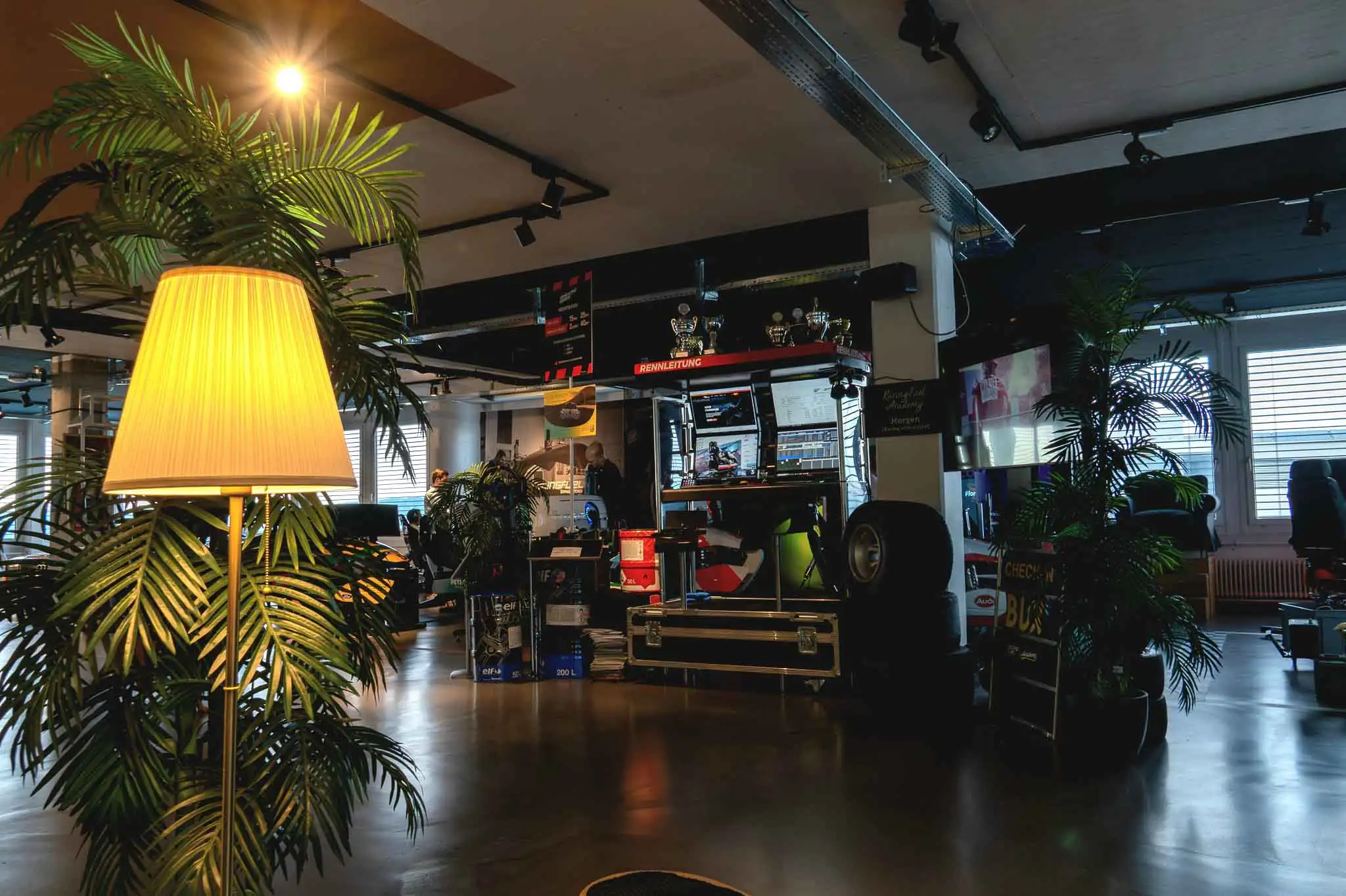Indoor racing team workspace with monitors, tires, trophies, potted plants, and a lit floor lamp.