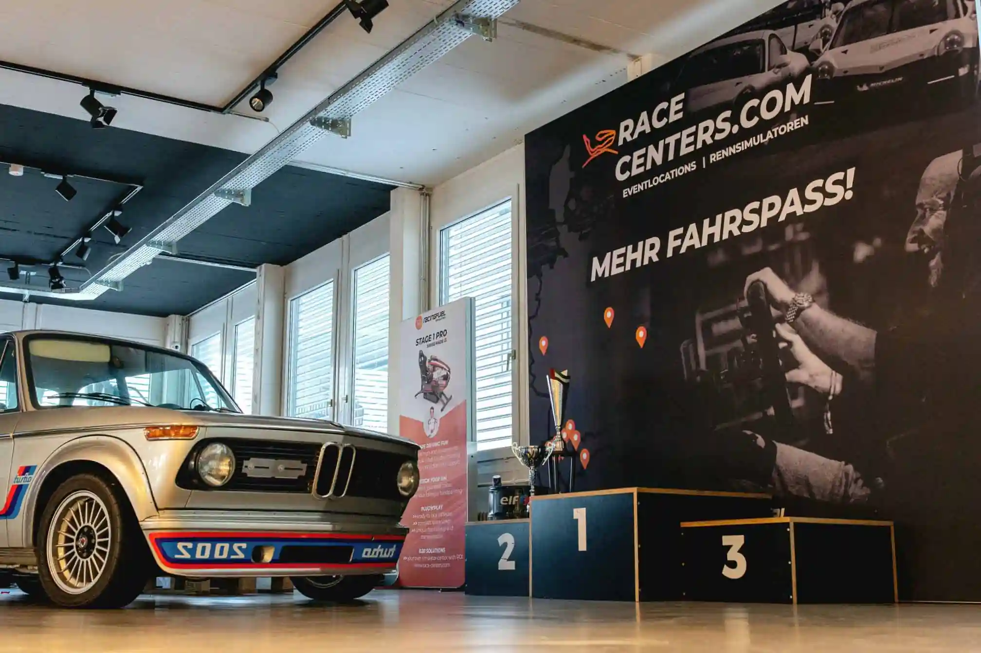 Classic silver BMW 2002 Turbo parked indoors next to a winners podium and a large racecenters.com advertisement banner.