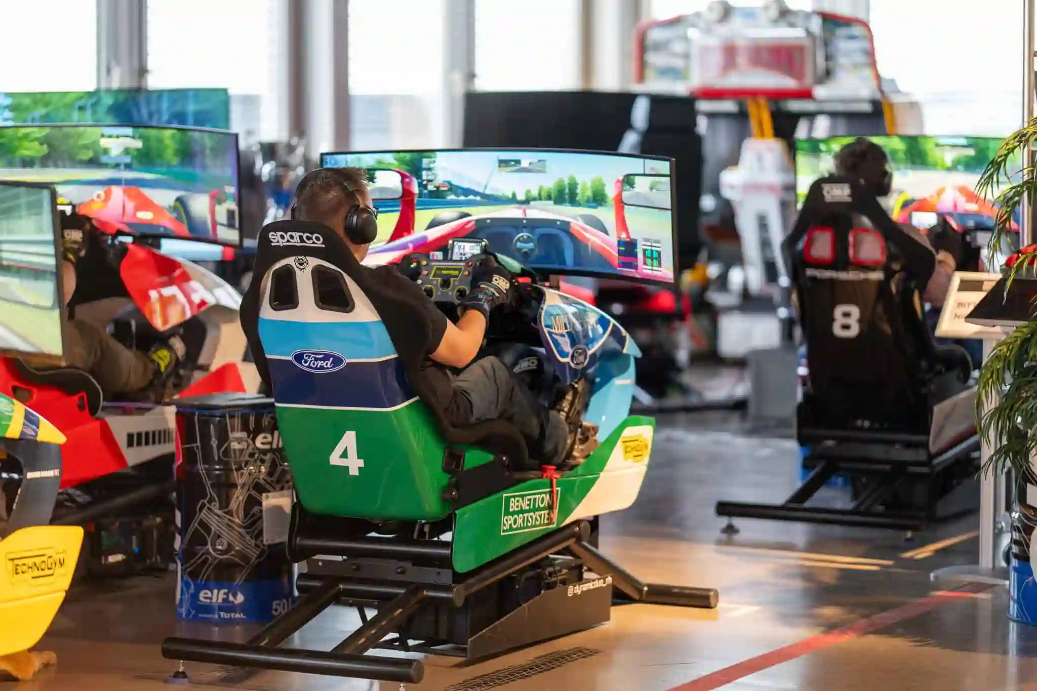 People seated in racing simulator pods with curved screens, steering wheels, and racing gloves, engaged in a virtual race.