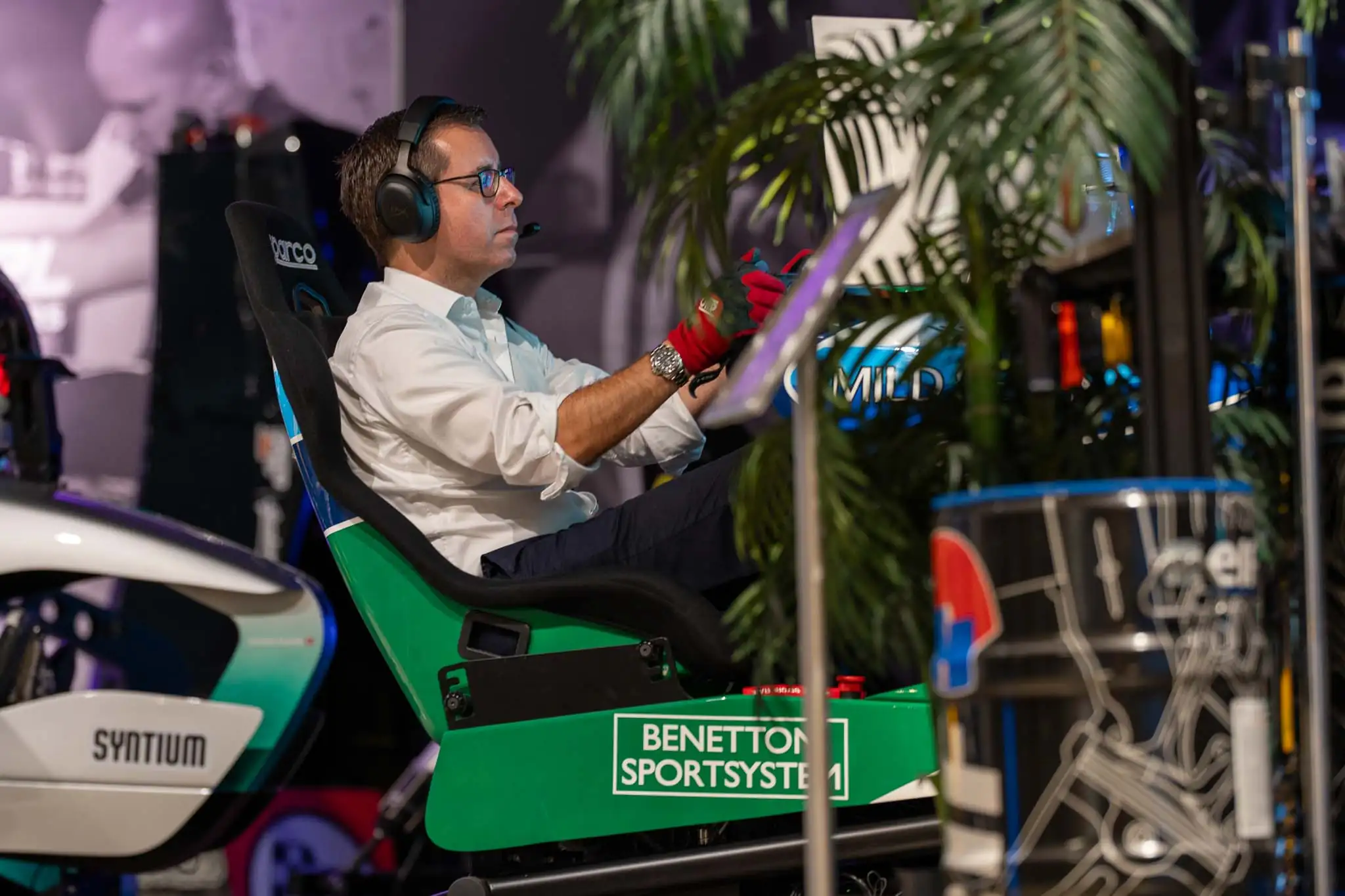 Man wearing headphones and red gloves sitting in a green Benetton SportSystem racing simulator seat.