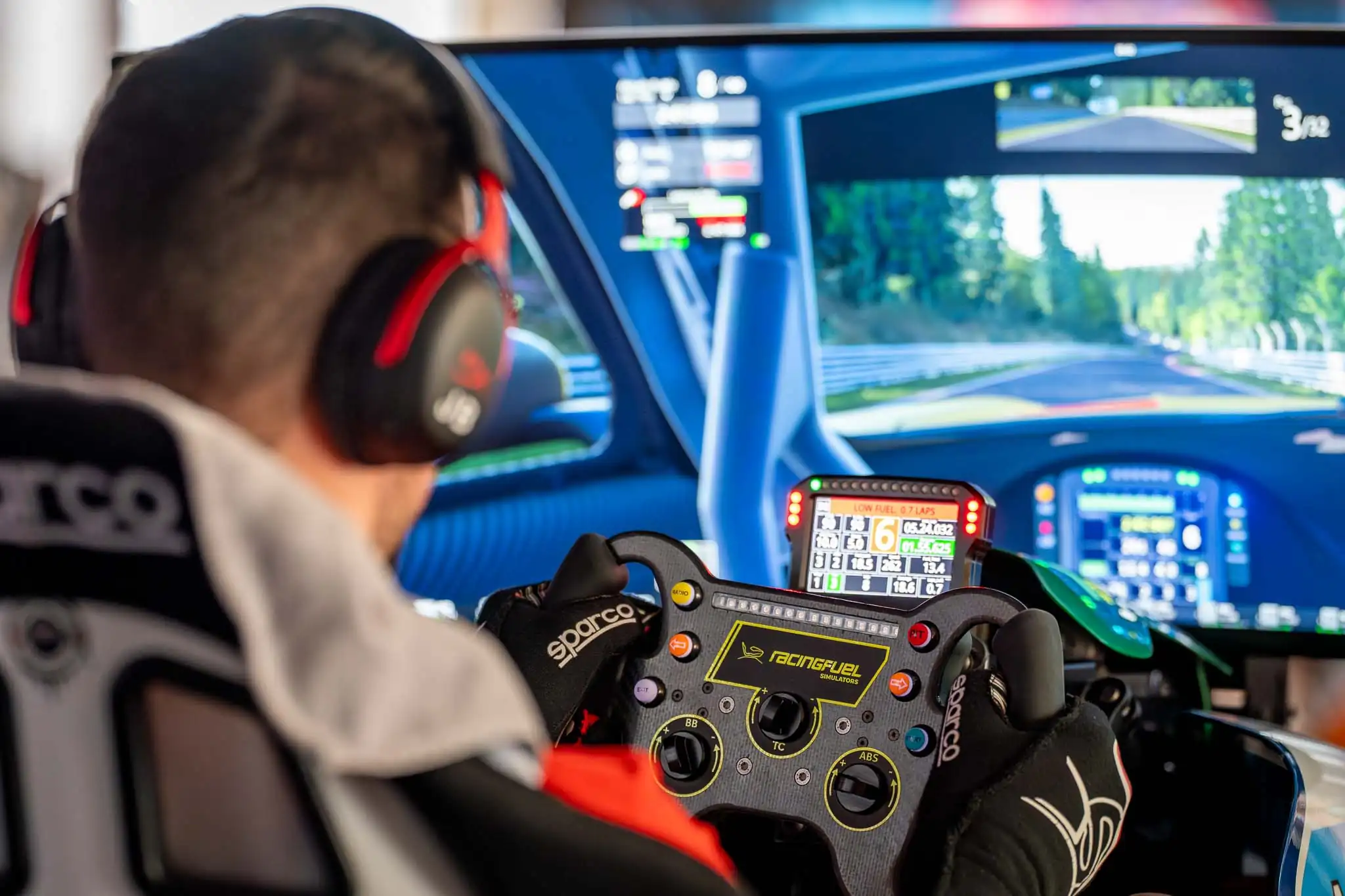 Person wearing headphones and racing gloves operating a RacingFuel simulator steering wheel with race track displayed on multiple screens.