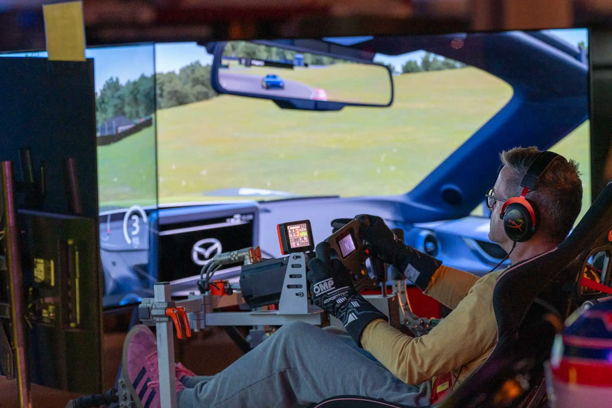Person wearing racing gloves and headphones using a driving simulator with a triple screen setup showing a racing game.