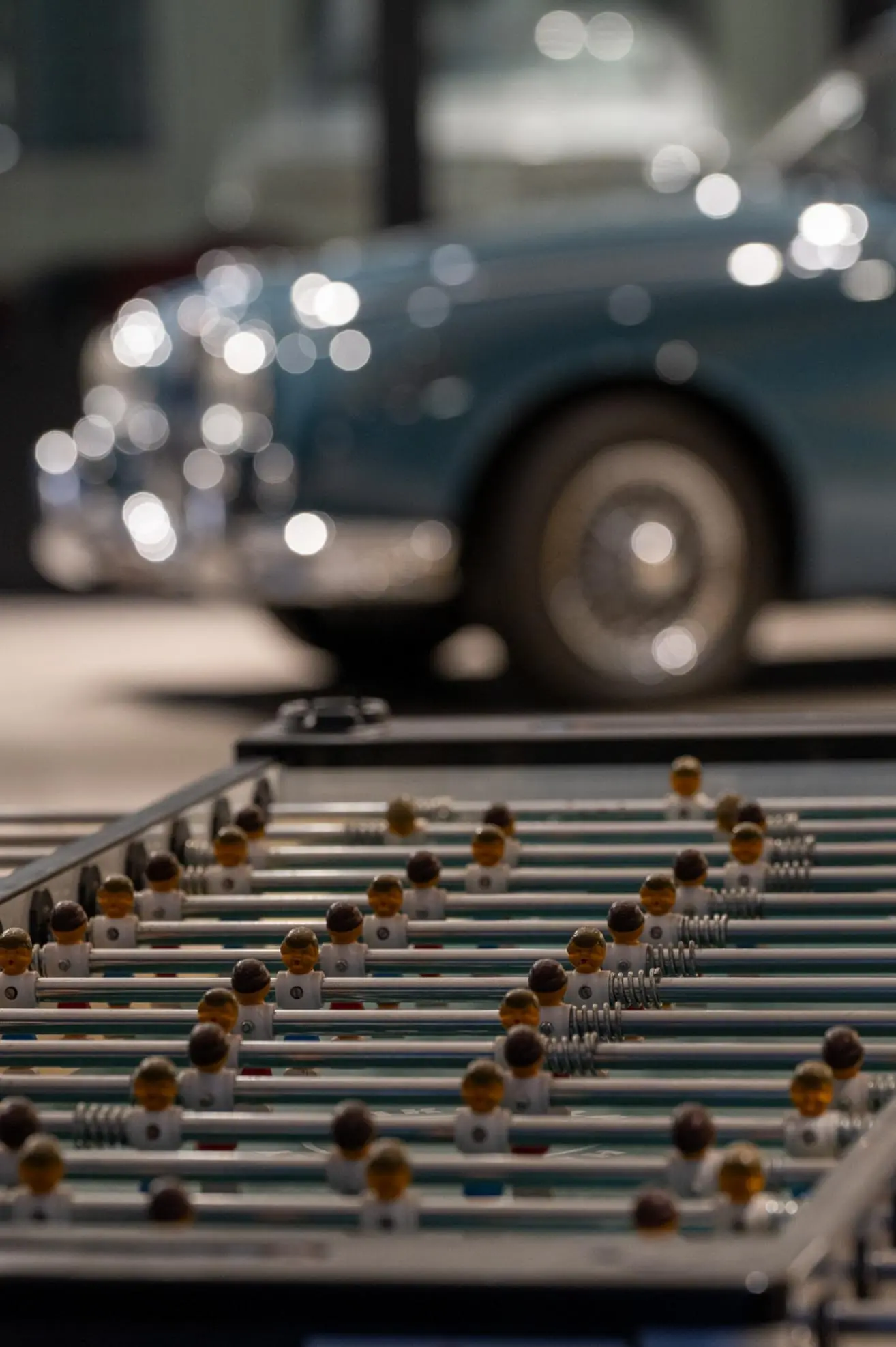 Foosball table with player figures in focus and a blurred vintage blue car in the background.