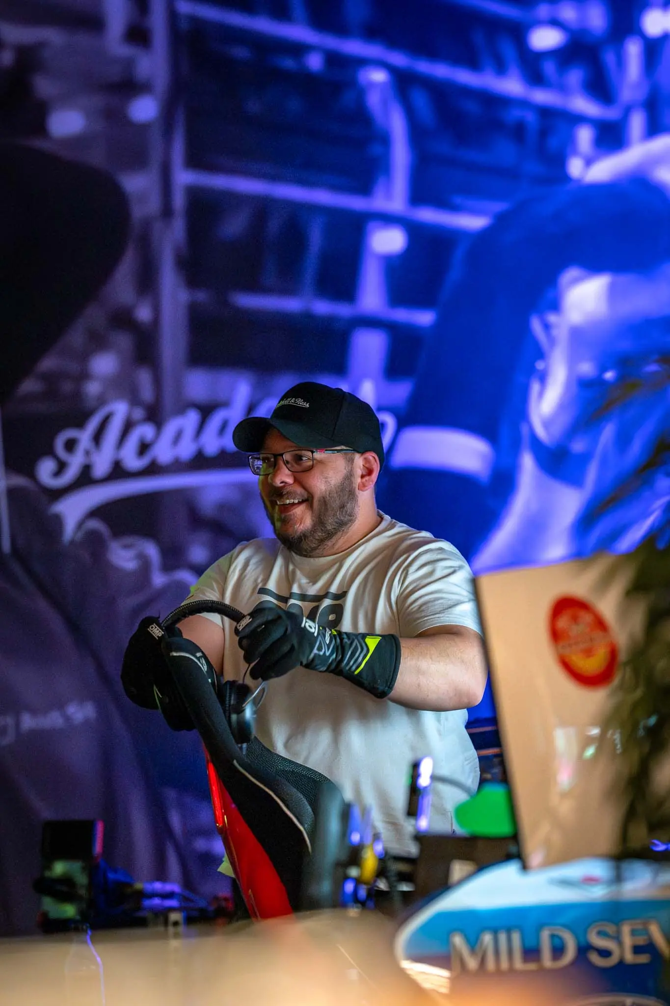 Smiling man wearing glasses, a black cap, and gloves adjusting a red and black chair indoors.