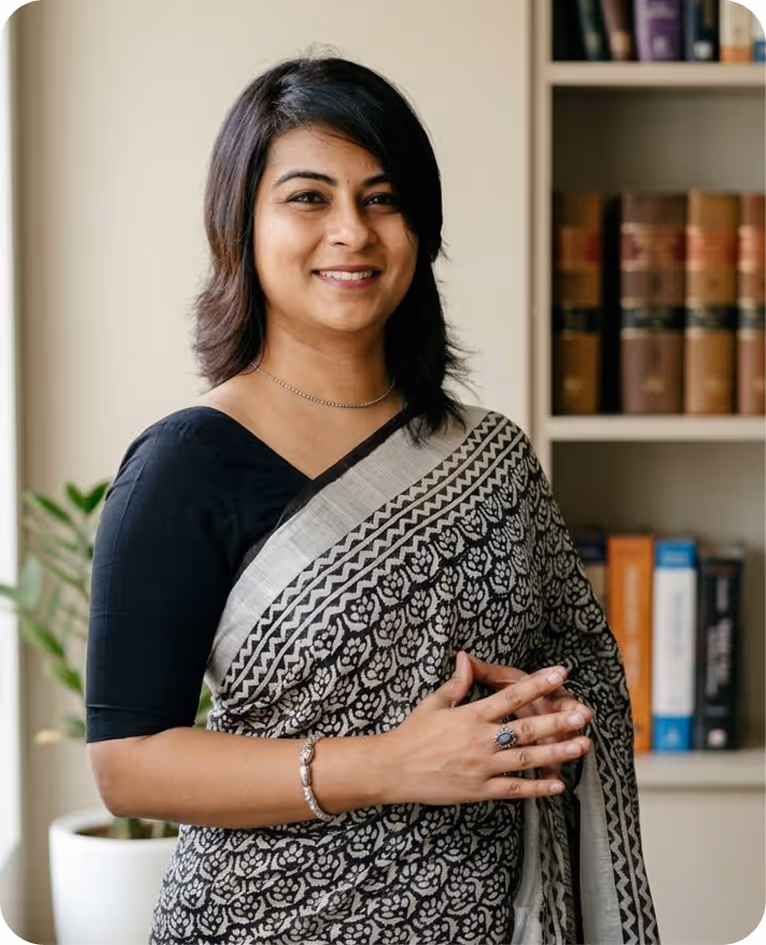 Smiling woman wearing a black and white patterned saree, standing indoors with bookshelves and a plant in the background.