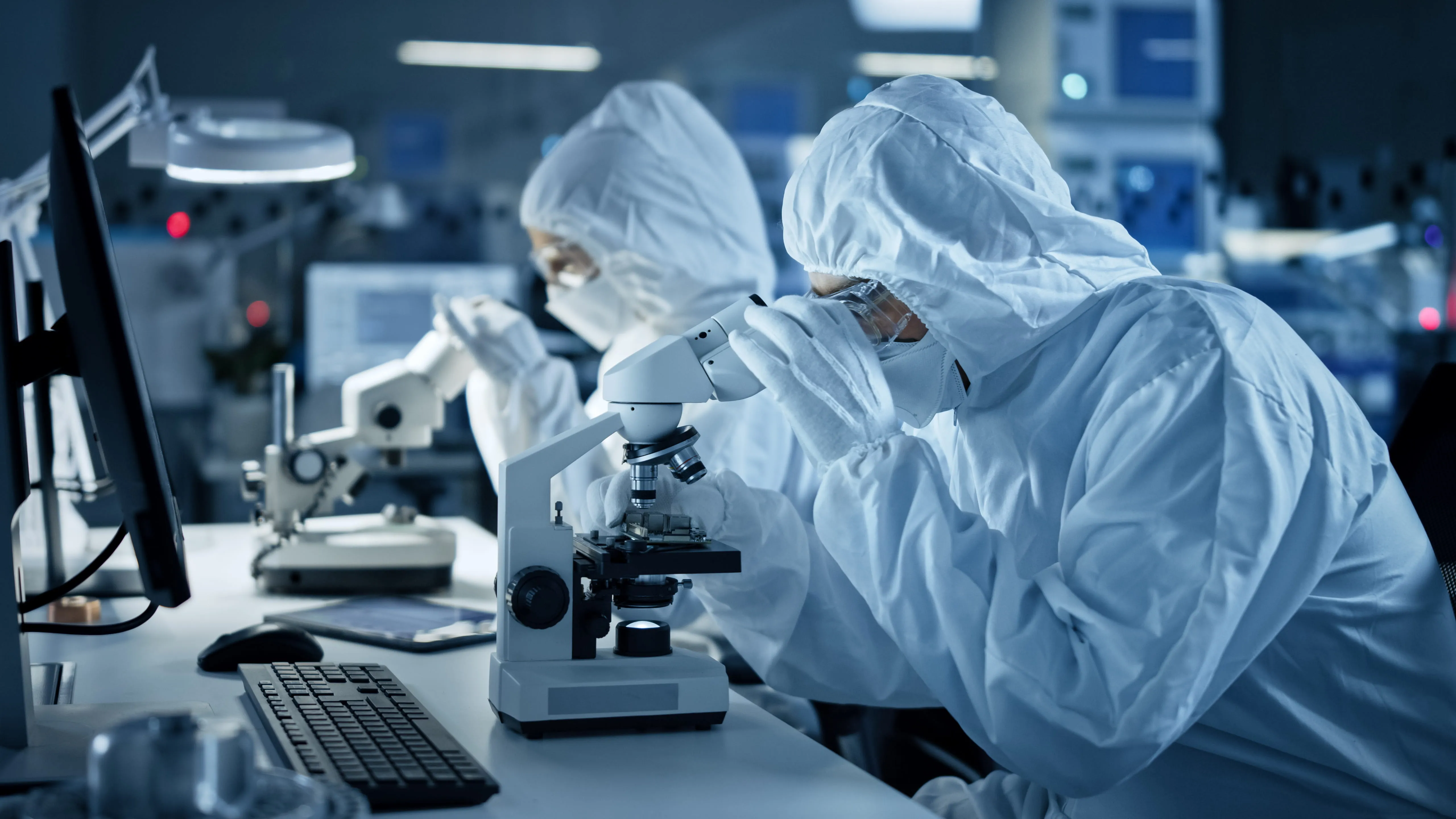 Two scientists in full protective suits looking through microscopes in a high-tech laboratory.