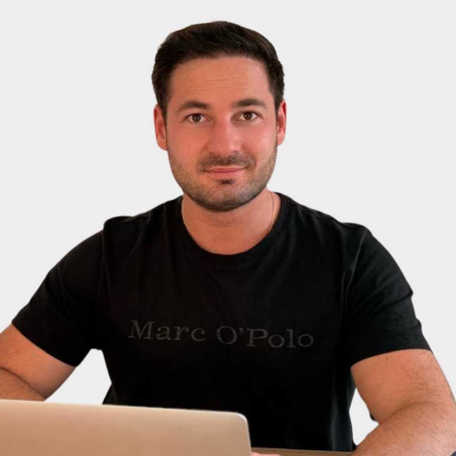 Man with short dark hair and beard wearing a black Marc O'Polo t-shirt sitting at a desk with a laptop.