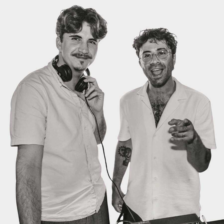 Two smiling men standing next to DJ equipment, one wearing headphones around his neck and the other pointing forward.