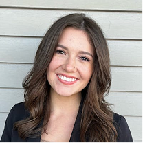 Smiling woman with long brown hair wearing a black blazer standing in front of a beige wooden wall.