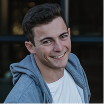 Young man with dark hair smiling, wearing a white shirt and a gray hoodie.