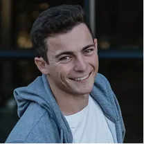 Young man with dark hair smiling, wearing a white shirt and a gray hoodie.
