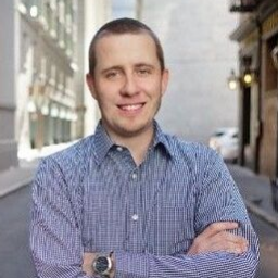 Smiling man with short hair wearing a blue checkered shirt standing outdoors with arms crossed.