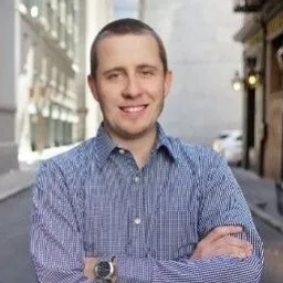 Smiling man with short hair wearing a blue checkered shirt standing outdoors with arms crossed.