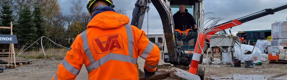 Construction worker in orange safety jacket and helmet operating an excavator on a muddy worksite with construction materials and equipment nearby.