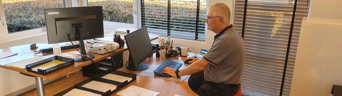 Older man with short gray hair sitting at a desk using a computer in an office with large windows and blinds.
