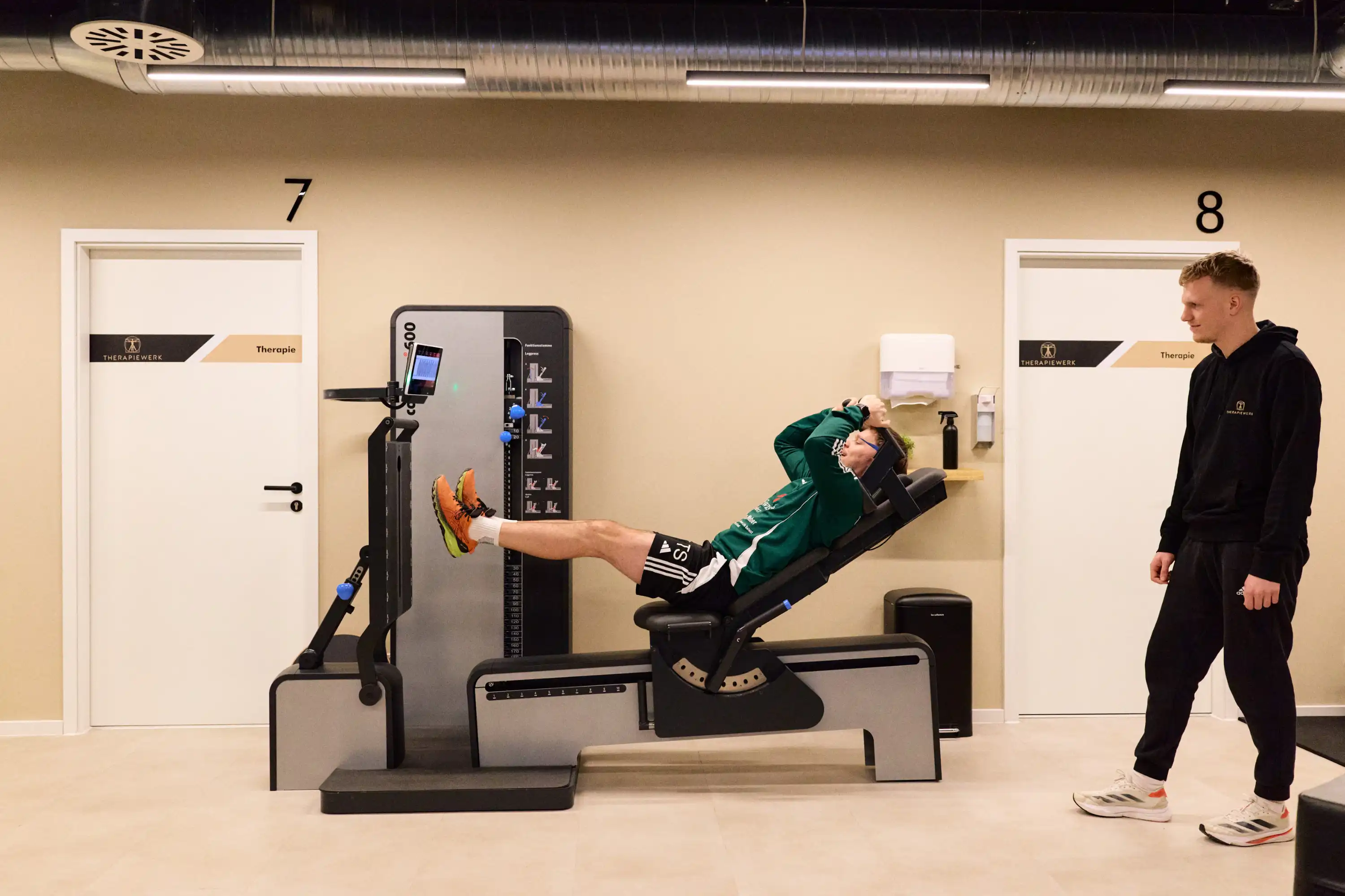 Mann in Grün trainiert auf Fitnessgerät, ein Trainer beobachtet ihn in einem Therapiezentrum.