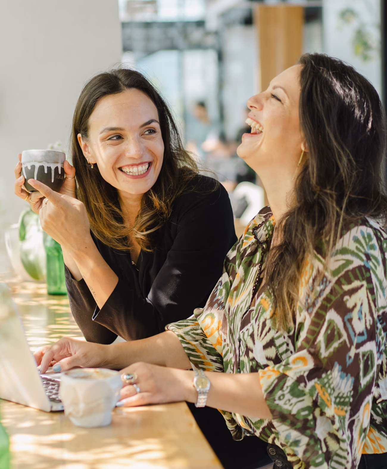 Two women sitting side by side at a table, smiling and laughing, one holding a cup and the other using a laptop.